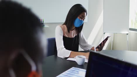 Asiatische-Frau-Mit-Gesichtsmaske-Und-Digitalem-Tablet,-Während-Sie-Auf-Ihrem-Schreibtisch-Im-Modernen-Büro-Sitzt