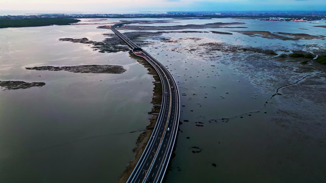 Drone captures sweeping views of Bali Mandara Toll road bridge crossing muddy low tide terrain framed by tropical vegetation and coastal atmosphere highlighting Indonesian infrastructure