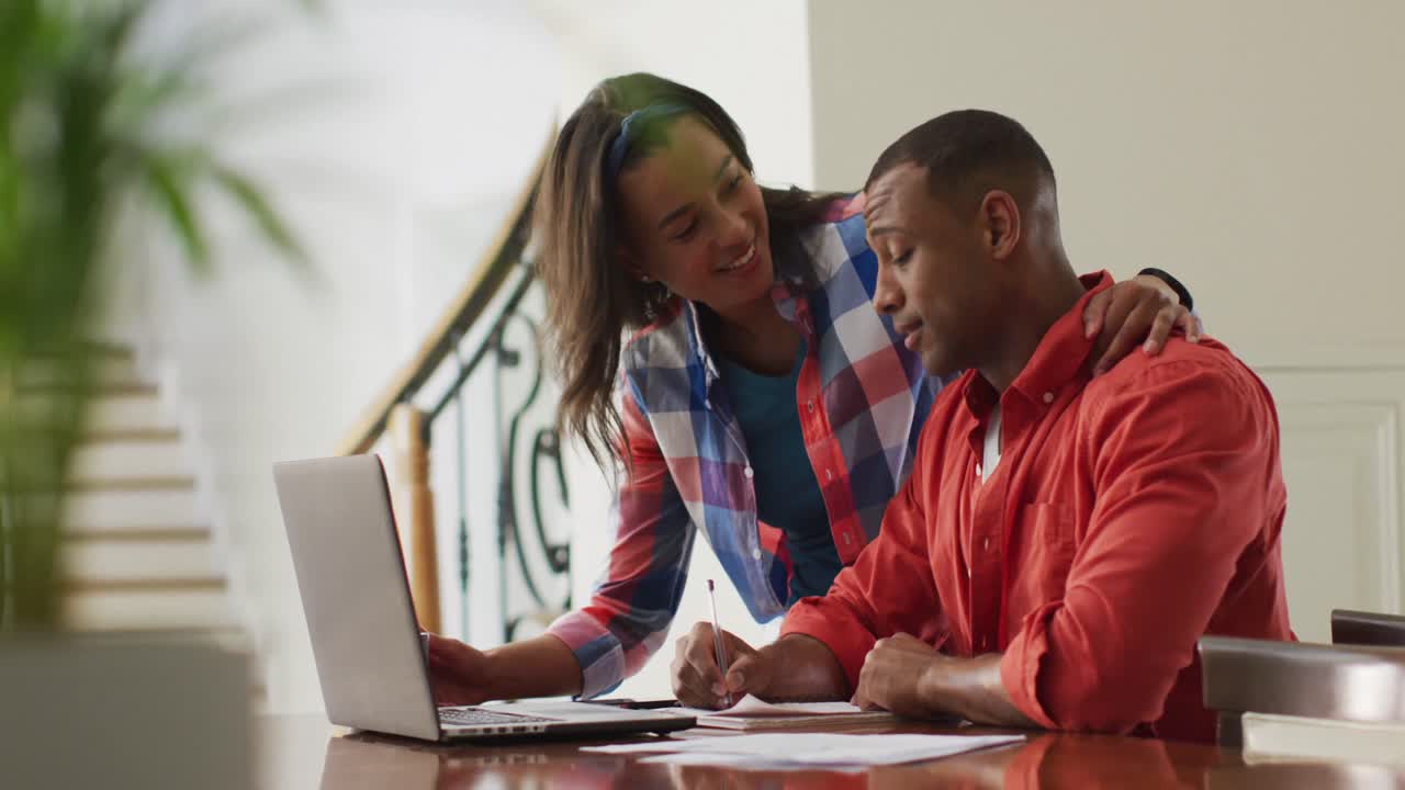 Happy biracial woman with coffee embracing partner sitting at desk with laptop and paying bills