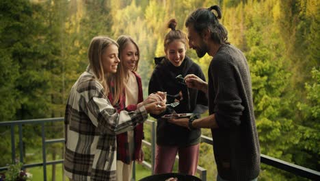 a group of friends try the food they cooked on the grill in a country house overlooking the mountains with a coniferous forest. the guy holds a plate of food and stands near the barbecue