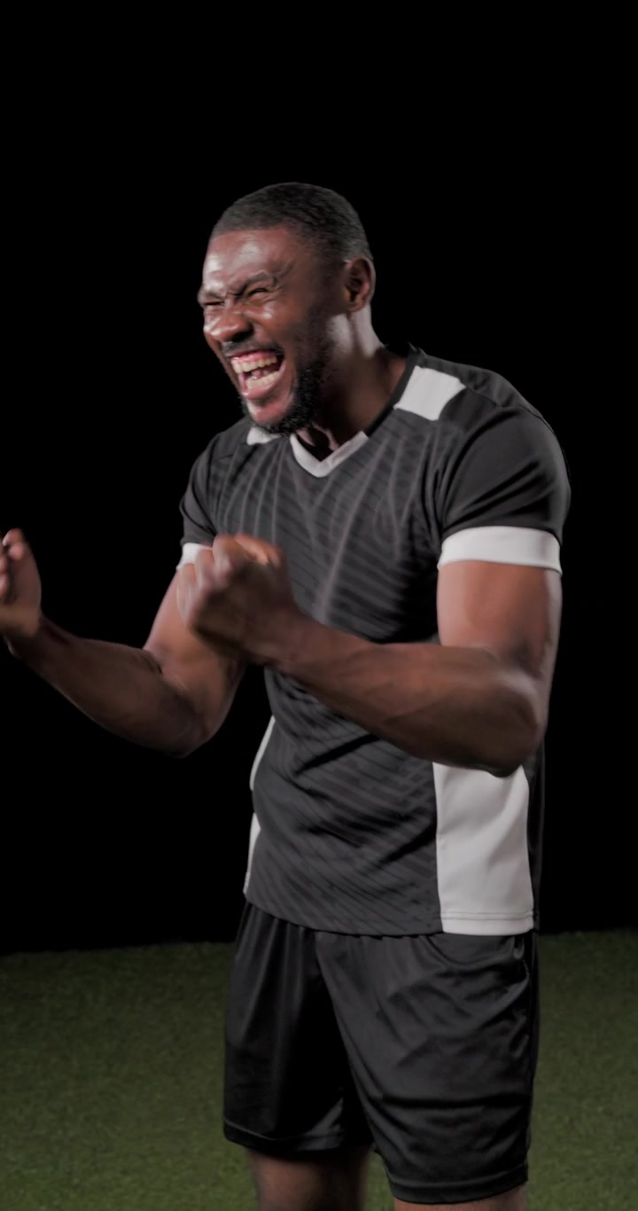 Uomo che entra nell'inquadratura, agita i pugni e indica, festeggiando su un campo sportivo in studio con un kit sportivo nero.