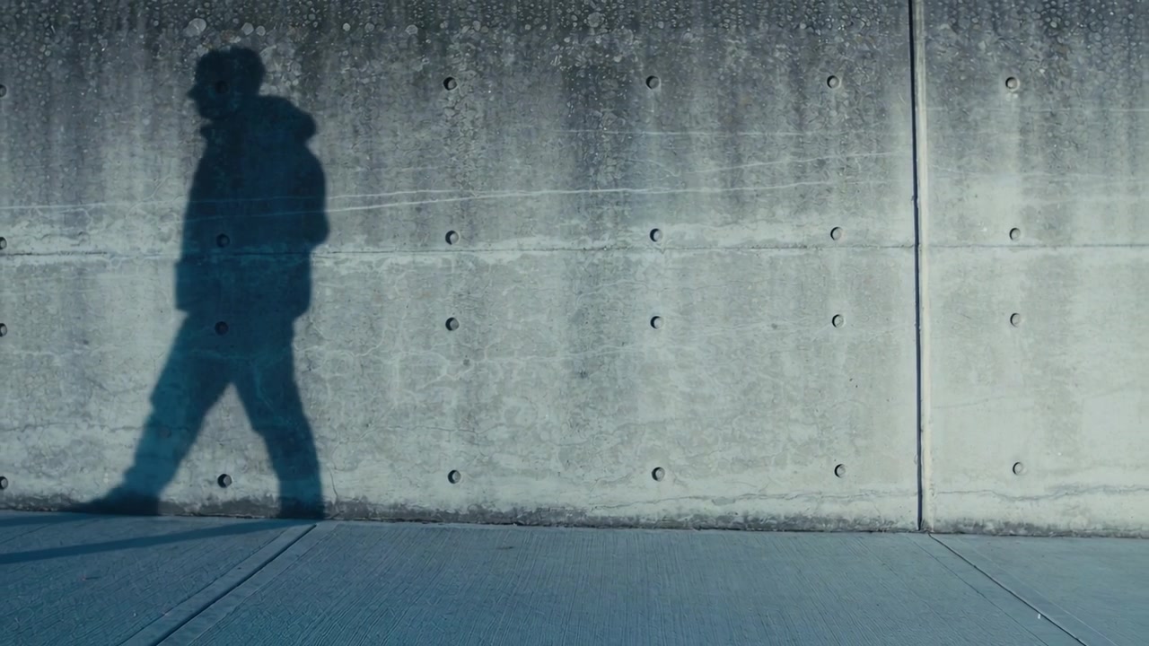 Walking hooded walker passing sidewalk while low sun casting long shadow on wall with rivet marks
