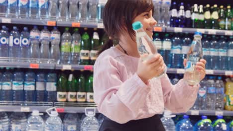 Close up video of a cheerful girl holding in her hands two bottles of water and dancing in the store over the bottles on the shelves background.