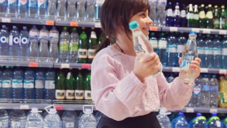 close up video of a cheerful girl holding in her hands two bottles of water and dancing in the store over the bottles on the shelves background.