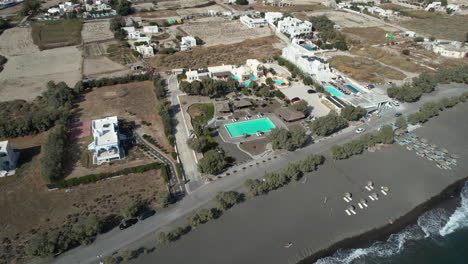 Vista-Aérea-De-Piscinas-Y-Casas-De-Verano-Junto-A-La-Playa-De-Arena-Negra-De-La-Isla-De-Santorini,-Grecia