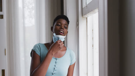 african american woman removing face mask and looking out of window at home