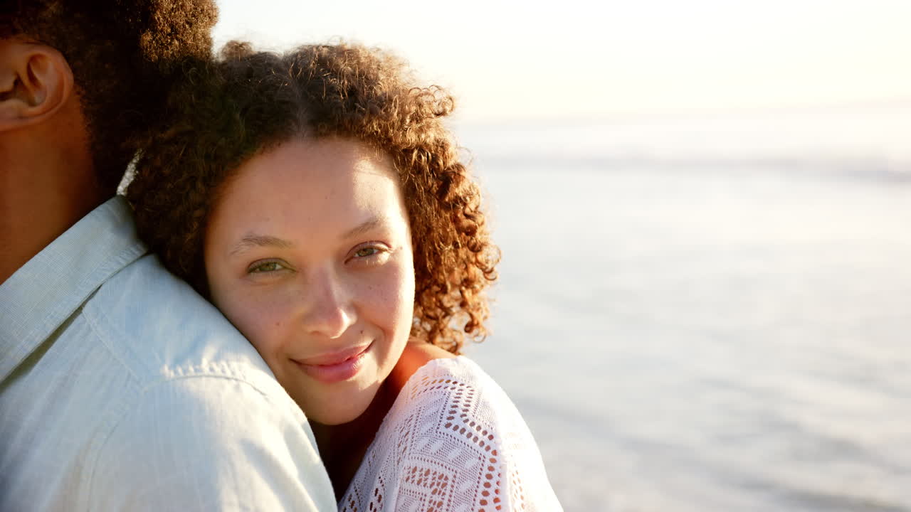 pareja biracial se abraza, mujer con cabello rizado sonriendo en la playa durante la puesta de sol con espacio de copia