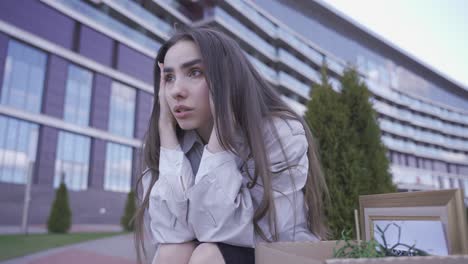 primer plano de una hermosa mujer sentada frente a un edificio de oficinas. ella tiene sus cosas en una caja. ella ha sido despedida. ella está triste.