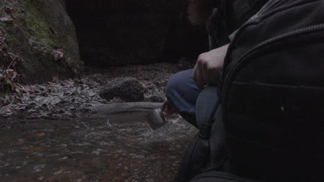 a man with a beard and a backpack scoops water from a stream with a tin cup after rinsing it out mossy boulders line the shore