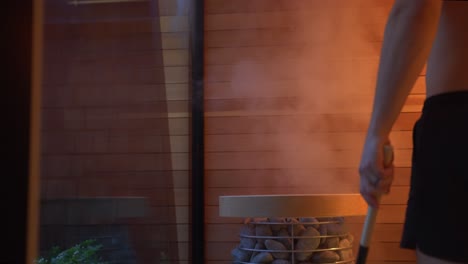 a man in a bathing suit stands inside a luxury wooden built sauna at night with an orange glow and pours water on the heater rocks with a large ladel and hot steam rises up to the roof