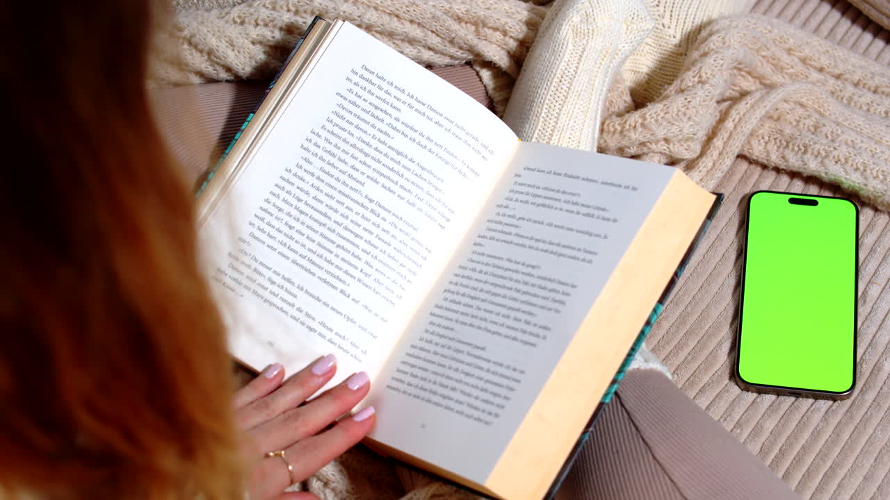 Female hands holding a book in a cozy setting, next to a smartphone with a green screen mockup. Ideal for reading, relaxation, or app mockup content, set on a soft blanket in a peaceful atmosphere.