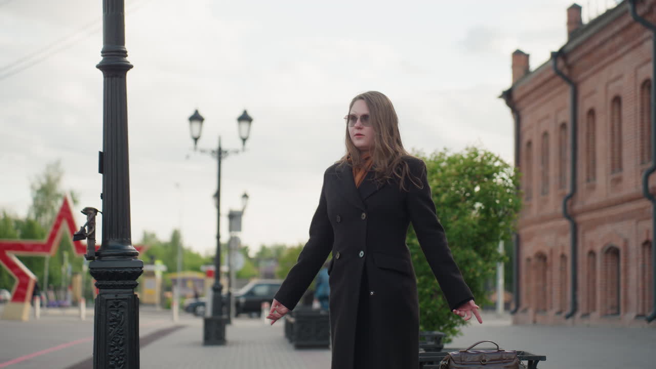 Man in dark suit and sunshade approaches woman sitting on bench near wooden art pillar partially blocking frame on city street lined with brick buildings creating cinematic urban depth