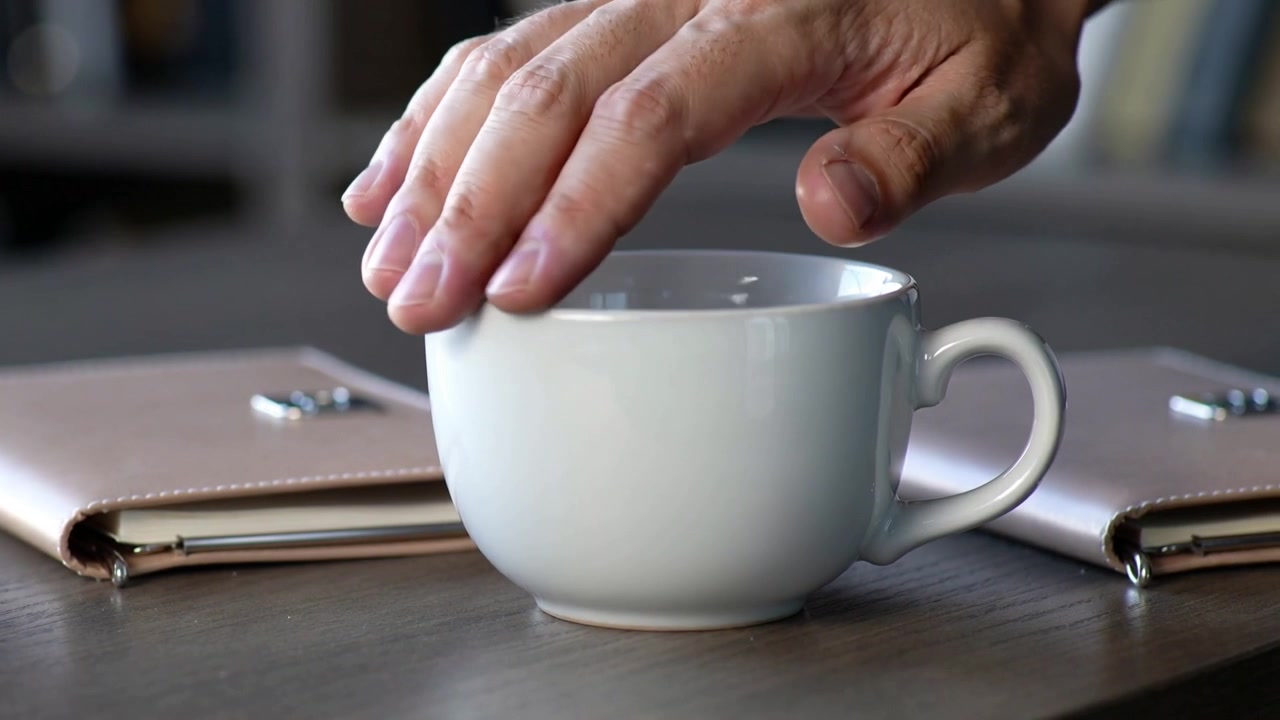 Entering right hand brushing rim of white mug on wooden table with tan notebooks and spoon