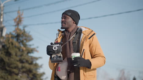low angle cameraperson holding drink in one hand holding camera in other while scanning city skyline under bright sun with blur power lines overhead on winter street with camera strap bag