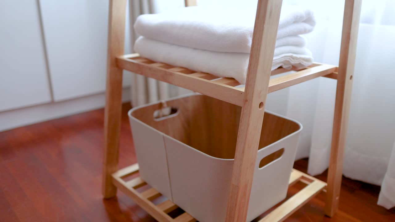 A person places a plastic laundry basket onto a wooden shelf holding folded white towels