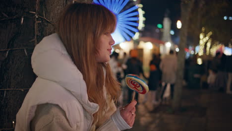 relaxed girl playing lollipop at christmas market night festive lights closeup.