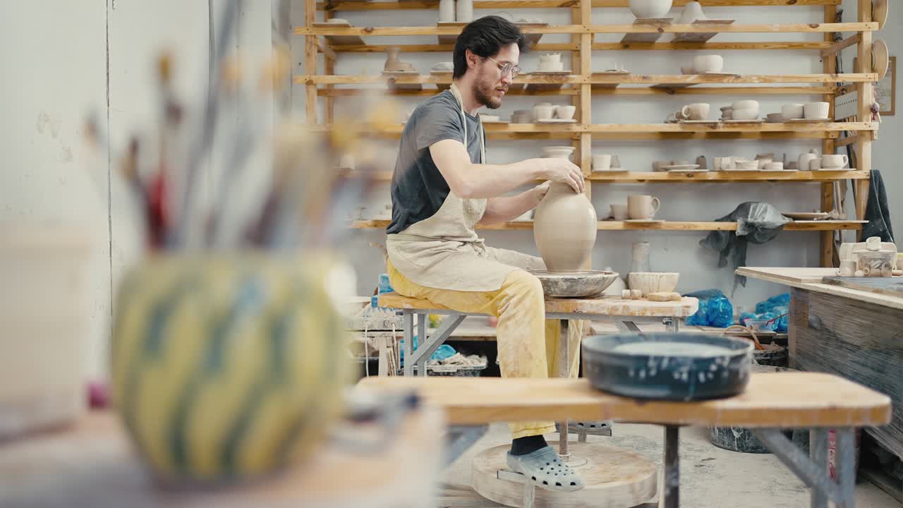 Male potter creating ceramic item on pottery wheel at workshop, focus moving from vase with brushes to artist