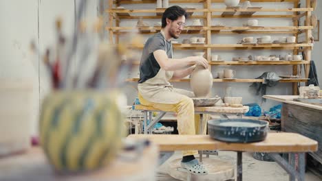 male potter creating ceramic item on pottery wheel at workshop, focus moving from vase with brushes to artist