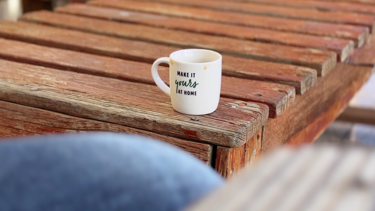 Una tazza in ceramica bianca con testo è posizionata su una panca di legno rustica e invecchiata all'aperto. La luce naturale soffusa e la profondità di campo ridotta creano un'atmosfera accogliente e di lifestyle per gli annunci sui social media.
