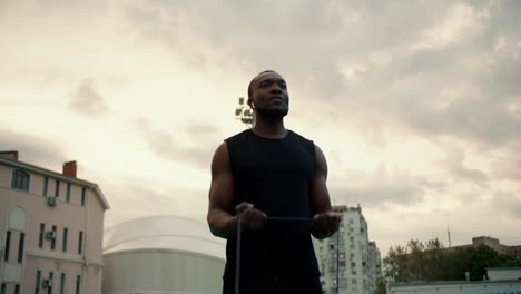 a black-skinned man in a black sports uniform trains using a special rubber simulator against a gray sky