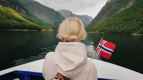 A-Woman-With-The-Flag-Of-Norway-Stands-On-The-Nose-Of-A-Cruise-Ship-Sails-On-A-Picturesque-Fjord-In-