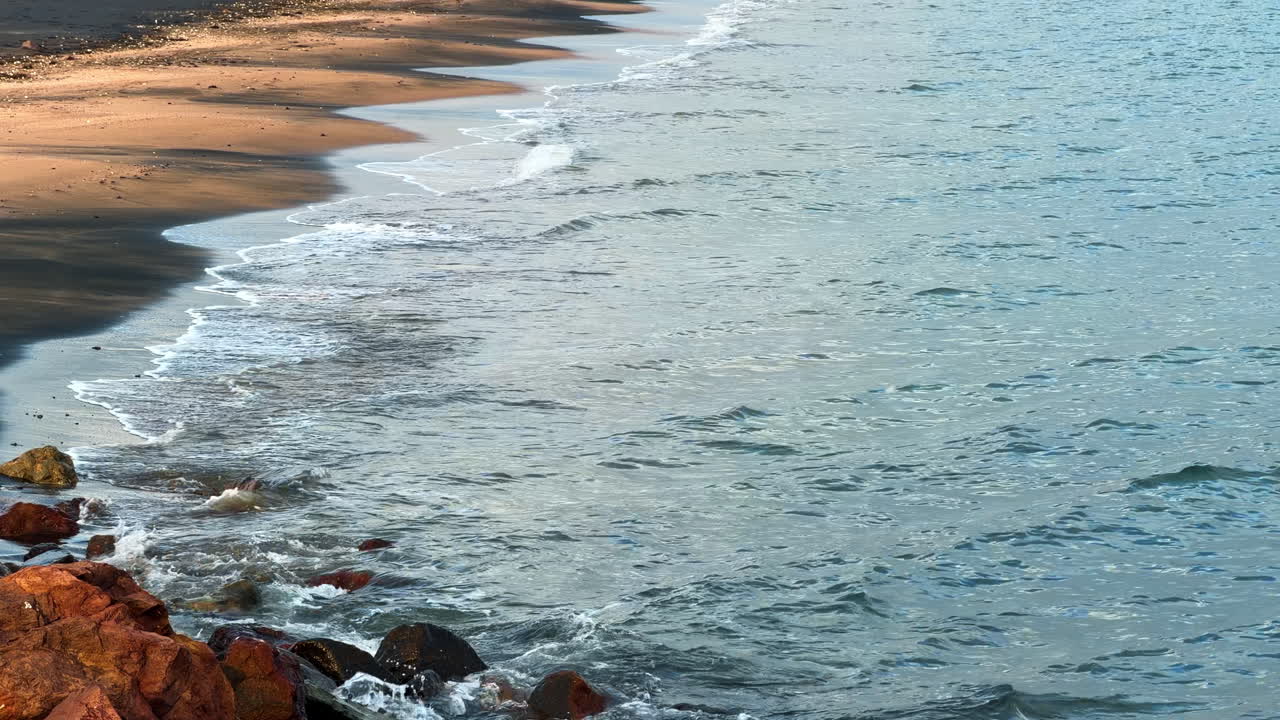 Le onde lambiscono dolcemente le rocce lungo la costa al tramonto. Il sole tramonta sull'acqua mentre le onde si infrangono e toccano le rocce sulla spiaggia. Colori tenui riempiono il cielo