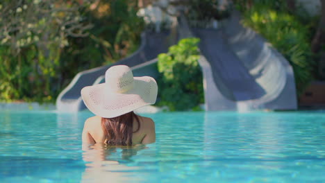 Frau,-Die-Im-Swimmingpool-Steht-Und-Einen-Großen-Hut-Trägt,-Der-Auf-Wasserrutschen-Blickt,-Die-Von-üppiger-Tropischer-Landschaft-Umgeben-Sind