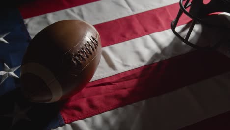 Studio-Shot-Of-Person-Picking-Up-American-Football-With-Helmet-On-Stars-And-Stripes-Flag-