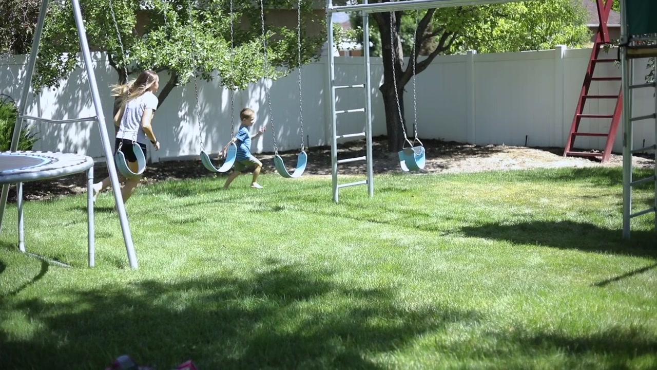 Slow Motion shot of a girl playfully chasing her younger brother around in the backyard by the playground.