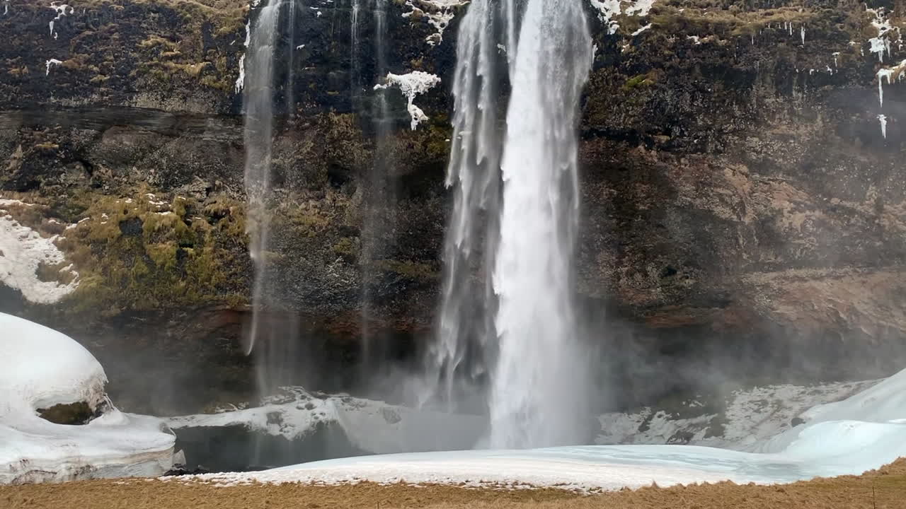 A waterfall cascades over a moss-covered cliff, surrounded by patches of snow and ice. Mist rises from the powerful stream, creating a dynamic contrast against the rugged rock face in Iceland