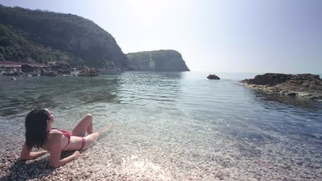 skinny lady in her red bikinis on the coast of montenegro on sunny summers days. lady sitting up in the water with small waves crashing against her body with the sun beating down playing with her hair
