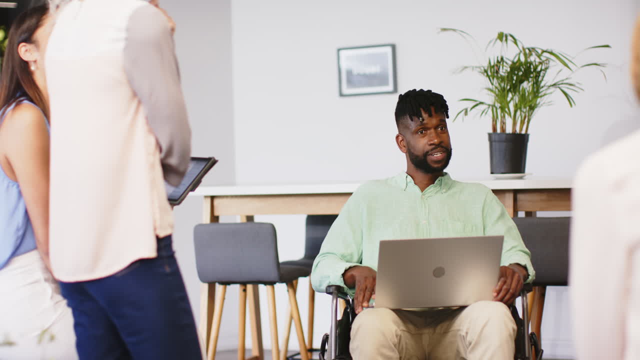 Smiling man in wheelchair using laptop, collaborating with colleagues in office