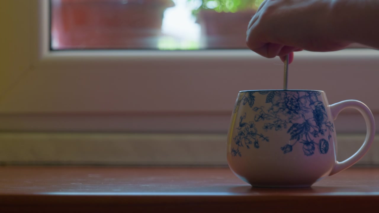 Hand roert met lepel in koffiekopje in een keramische mok met patroon op een houten vensterbank. Ochtendroutine ontbijtdrank, bereiding van hete thee met wazige raamachtergrond, statisch shot