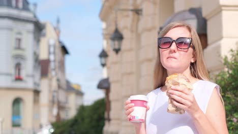 a beautiful girl eats a donut and drinks coffee standing the background of a beautiful building