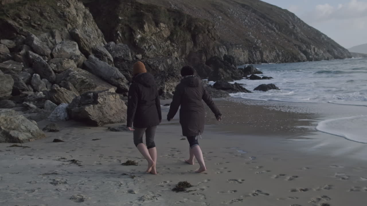 Two women in black coats walking barefoot on the beach. Handheld wide shot with slow motion. Waves coming in. Stony cliff in the background.