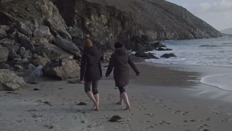 two women in black coats walking barefoot on the beach. handheld wide shot with slow motion. waves coming in. stony cliff in the background.
