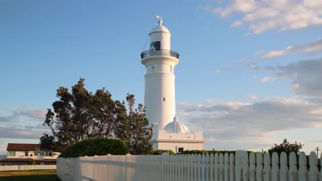 Dolly-shot-of-white-light-house-with-picket-fence-in-the-foreground-at-sunset