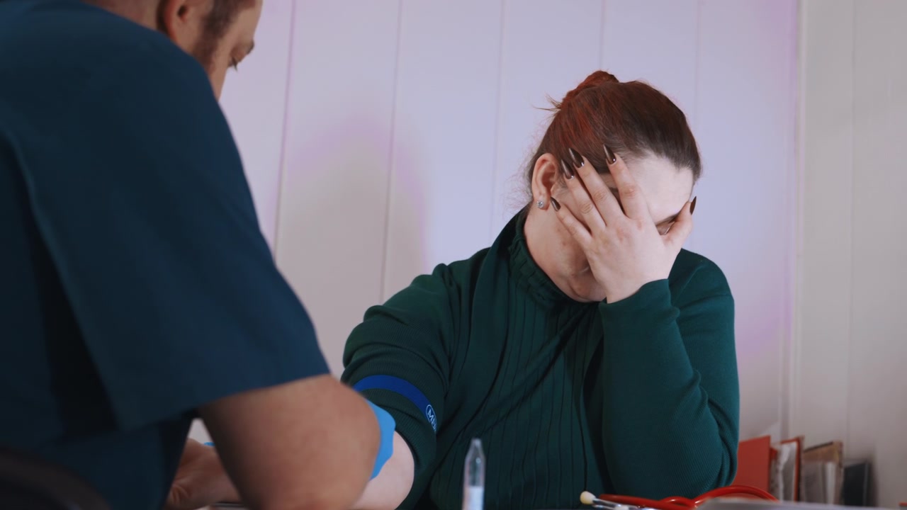 Paramedico che prepara un prelievo di sangue a domicilio, con laccio emostatico applicato mentre il paziente si copre il viso per l'apprensione, siringa e provette sul tavolo, procedura di flebotomia mirata in un contesto domestico.