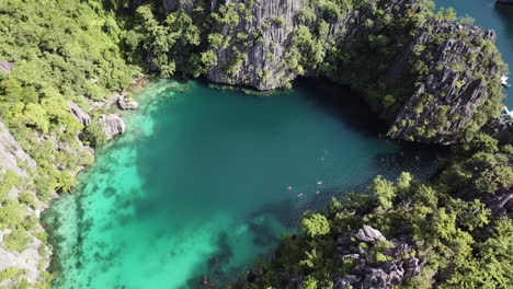 People swimming between high cliffs and behind a rocky insland in turquoise clear water towards the boats on a bright sunny summer day. Drone dolly and truck shot