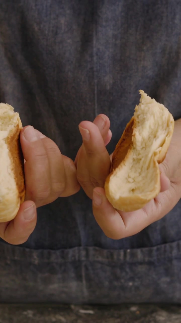 Vertical video: Tearing baker in denim apron revealing crumb on flour counter near rack, copy space