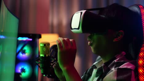 close up side view of asian boy streamer playing game, touching, and swiping experience virtual reality in futuristic goggles. green screen personal computer on desk illuminated by rgb led strip light