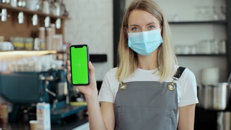 portrait of caucasian woman in mask standing behind bar and holding smartphone with chroma key. young female barista in madical mask looking at camera and showing green screen in coffee shop. business concept.