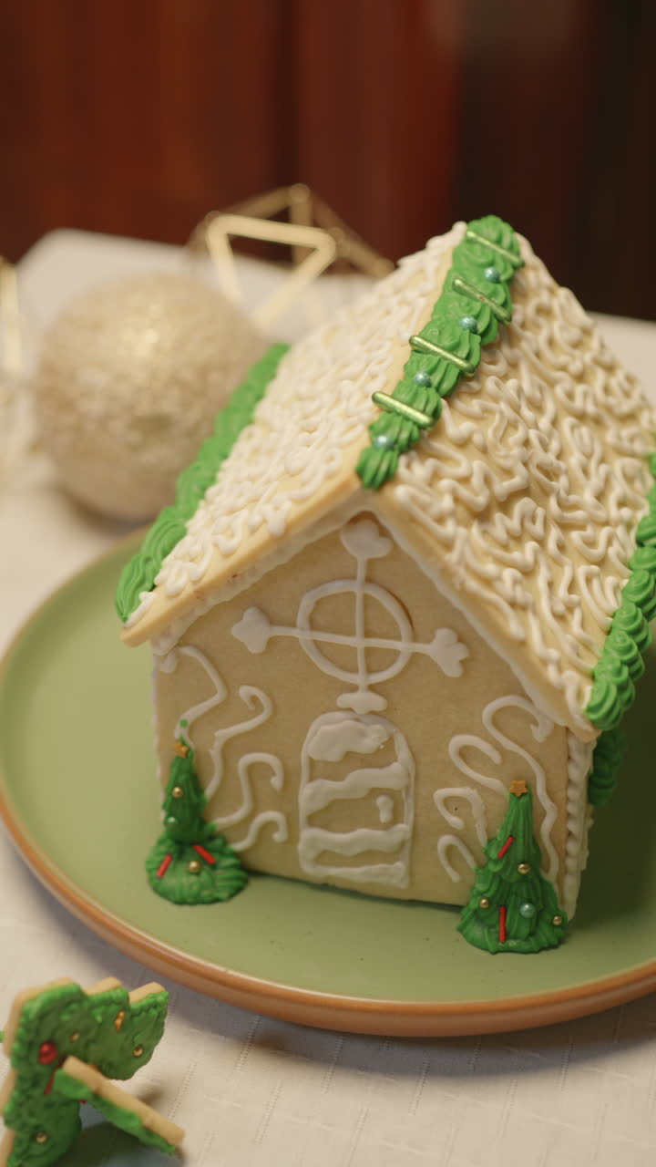 Close-up of a Christmas house made of cookie and decorated with detailed icing in white and green tones, with small edible trees and a warm atmosphere
