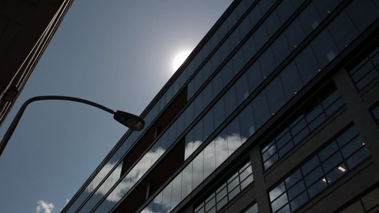 Vista dinámica de un edificio moderno con fachada de cristal que refleja el cielo y las nubes pasajeras. Mientras el sol brilla intensamente, creando un destello prominente detrás de su borde. Destacando la arquitectura urbana