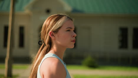 side view of young lady walking outdoors with hair tied back, looking thoughtful, with blurred background including building and greenery, sunlight illuminates scene while distant bird flies by