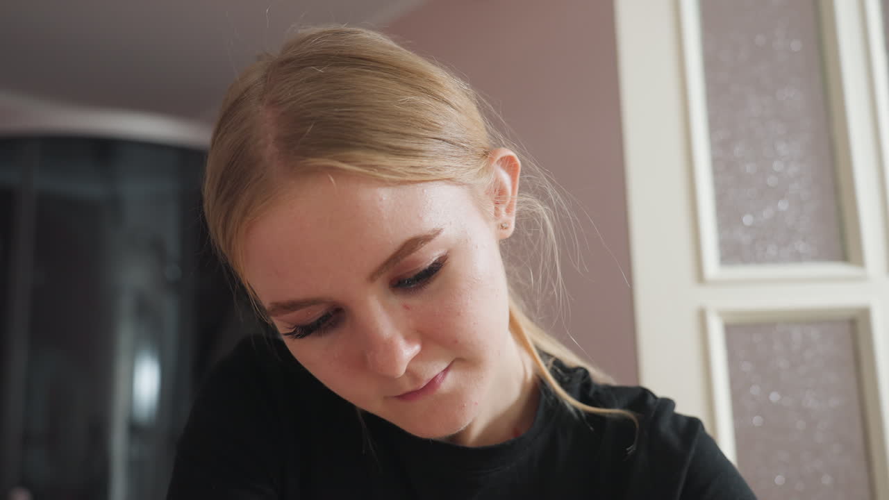 Light skin blonde woman with middle-parted hair tucked behind ears wearing black shirt, looking down thoughtfully in softly lit interior with blurred frosted glass door in background