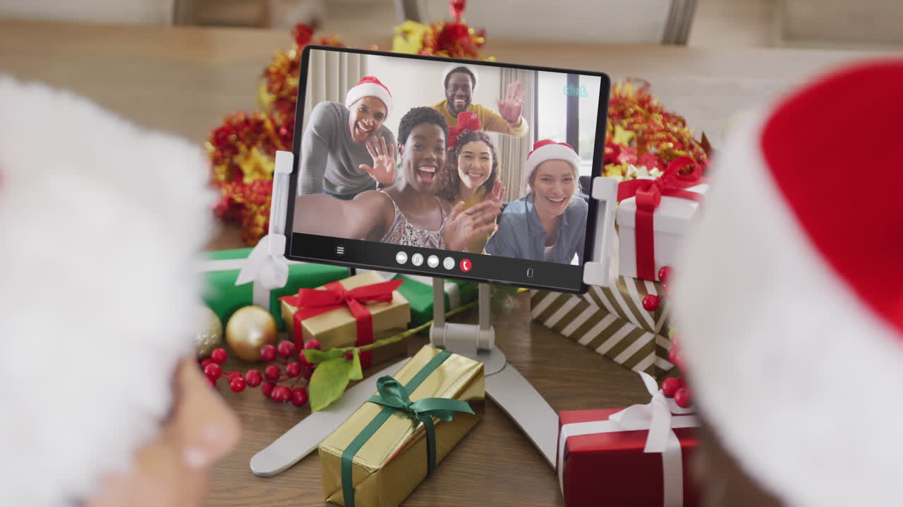 animación de una pareja diversa con sombreros de papá noel en una llamada de video de tableta con amigos en navidad