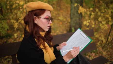 rear view of young lady in sunglasses, yellow beret, and yellow muffler deeply focused on her drawing, seated on a bench amidst vibrant autumn foliage