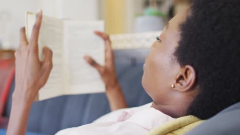 happy african american woman lying on sofa in living room, reading book