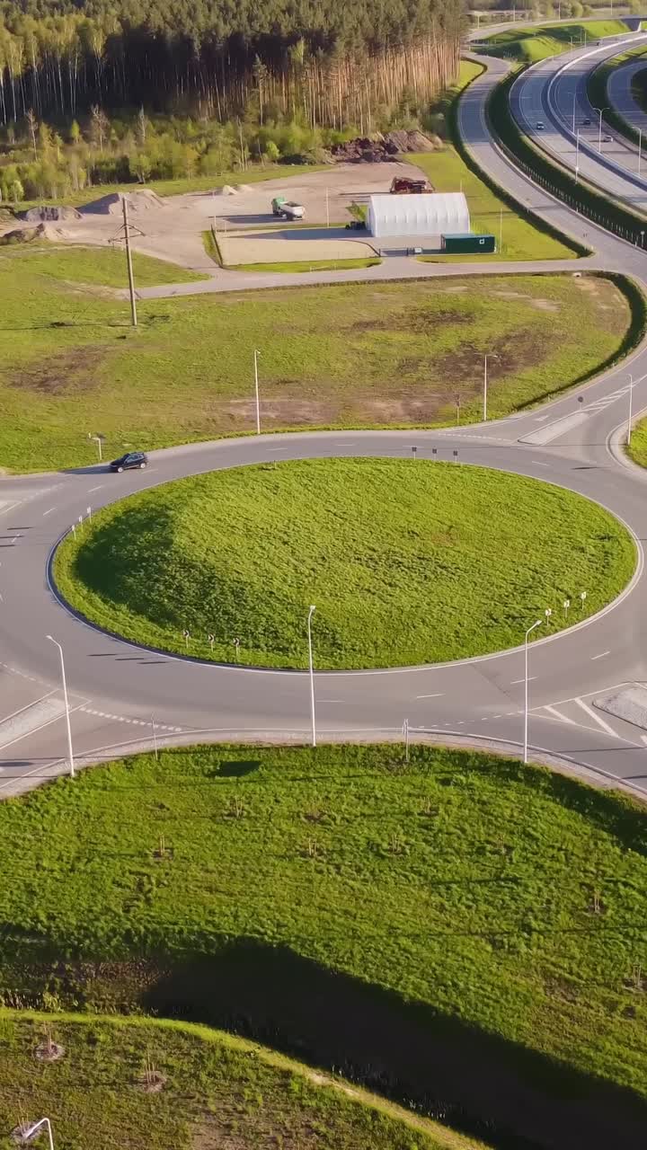 Vertical drone shot frames the Katlakalns roundabout with green mound in the center, nearby coniferous pine forest, and industrial sheds with materials visible along the access road, orbiting aerial